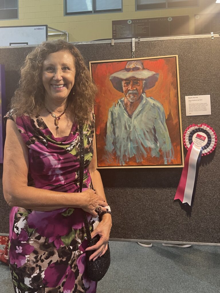 The Dayboro Art Gallery is buzzing with activity. 3 Artist standing beside her winning Portrait with a vibrant orange background